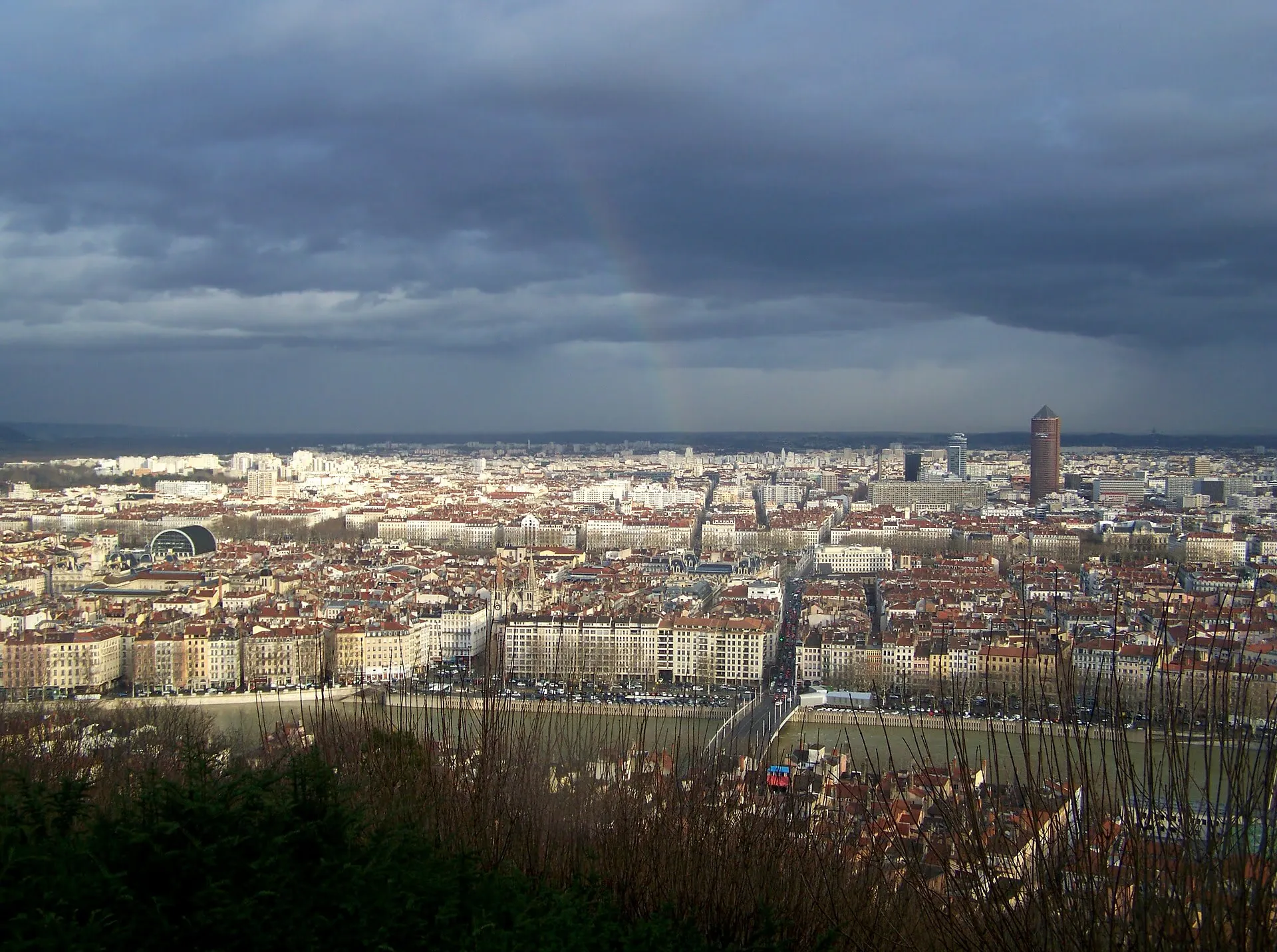 Vue panoramique de Lyon depuis la colline de Fourvière, sous un ciel gris chargé d'averses, avec les toits de la ville à perte de vue.