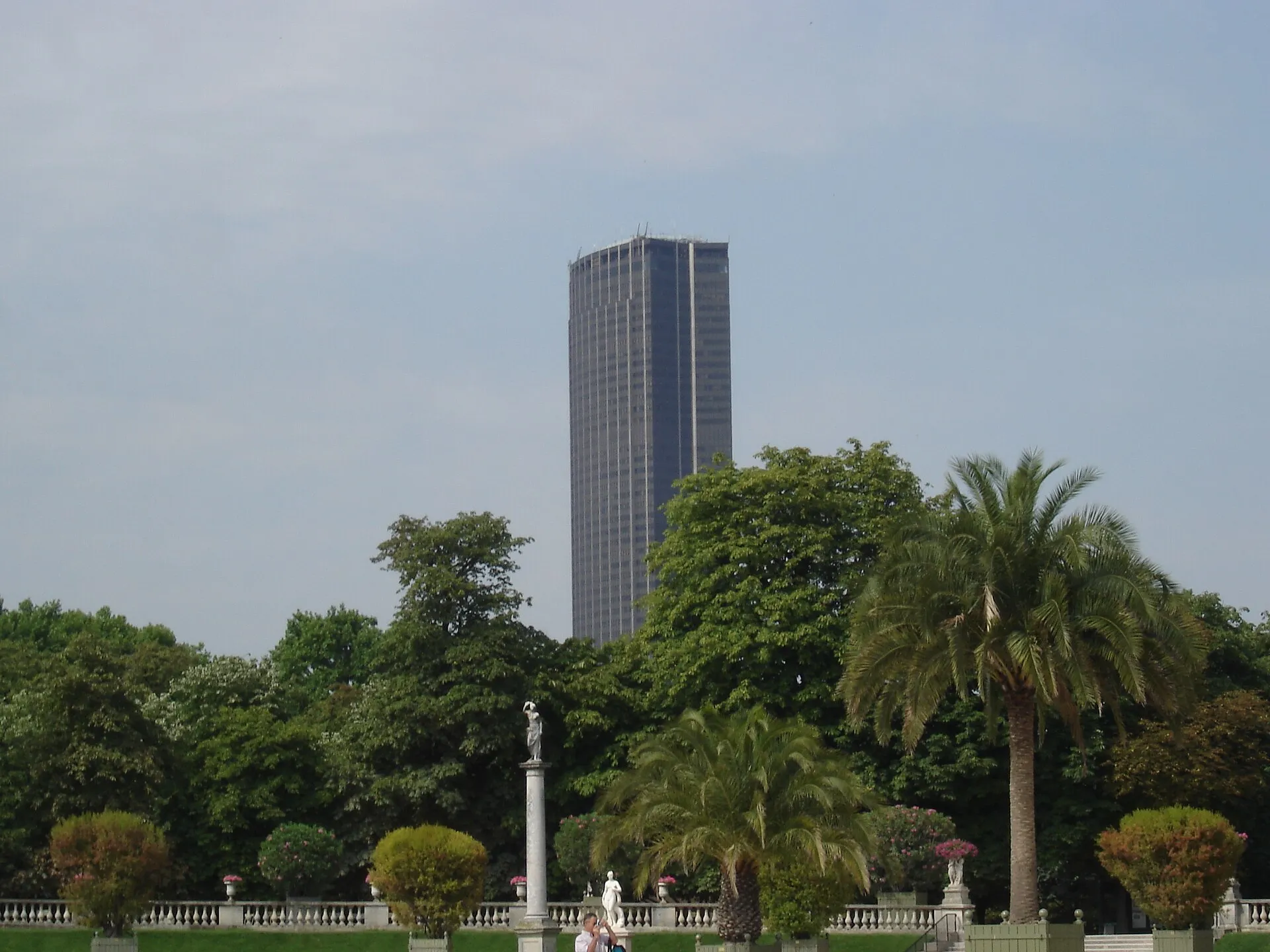 La Tour Montparnasse vue depuis le jardin du Luxembourg, dépassant des arbres et des palmiers du jardin.