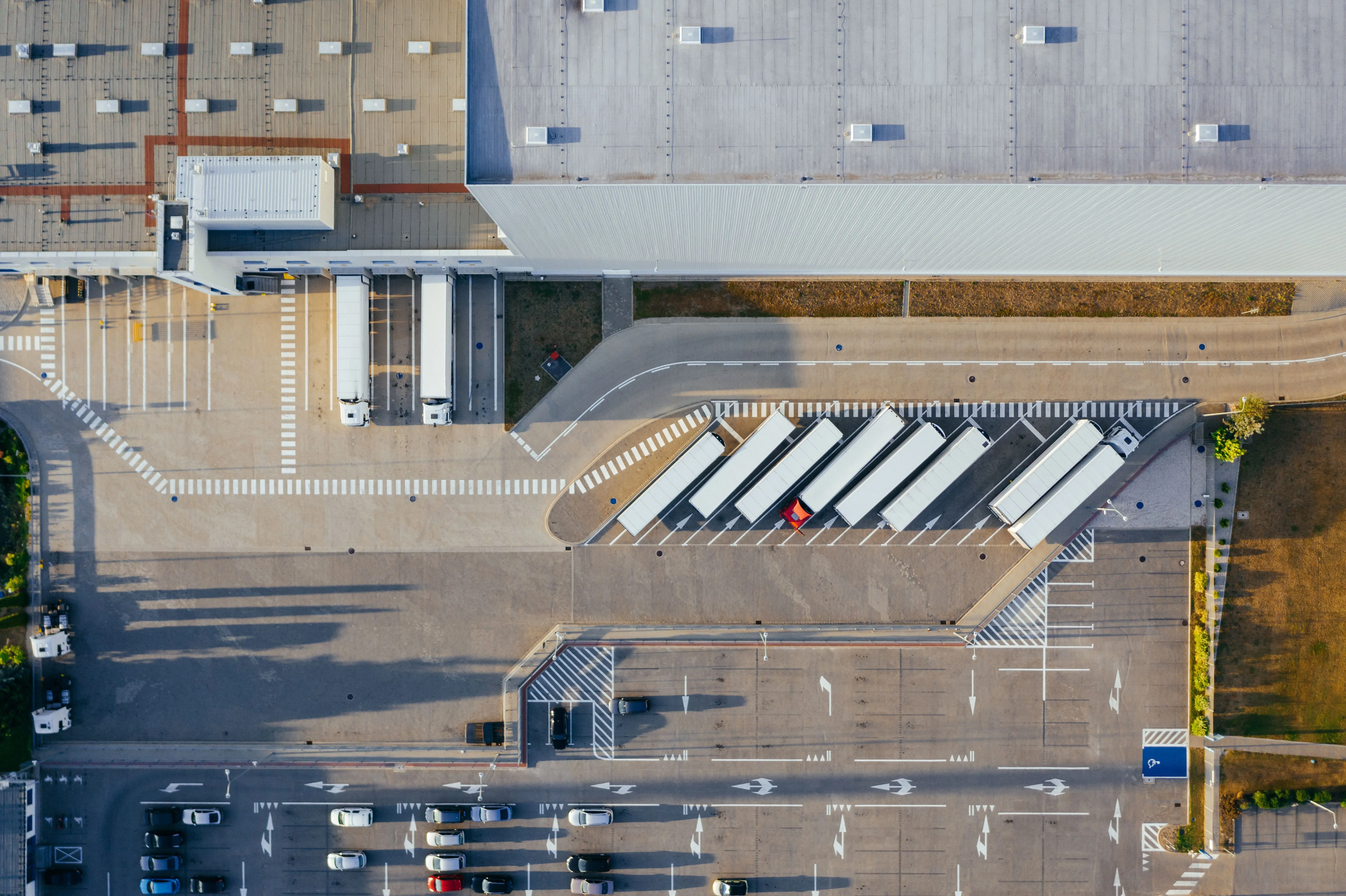 Vue aérienne d'un centre logistique, longs entrepôts à toiture claire alignés le long d'allées de circulation, camions et remorques stationnés sur les aires de manœuvre, champs et zones boisées autour